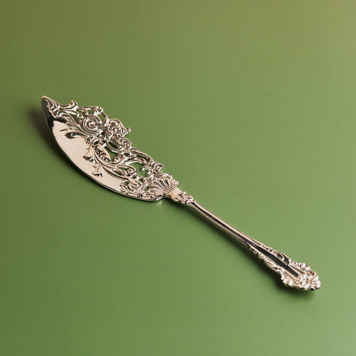 A handcrafted antique style brass cake server with intricate designs on the handle, placed against a green background.