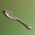 A handcrafted antique style brass cake server with intricate designs on the handle, placed against a green background.