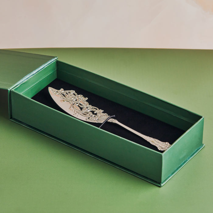 A handcrafted antique style brass cake server with intricate designs on the handle, placed against a green background.